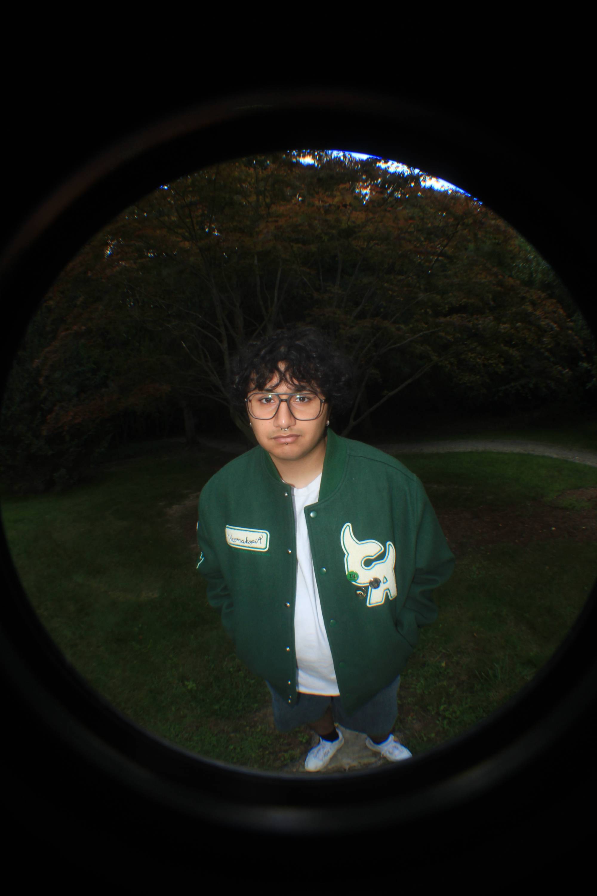 Young man with curly black hair and glasses wearing a dark green varsity jacket with trees in the background